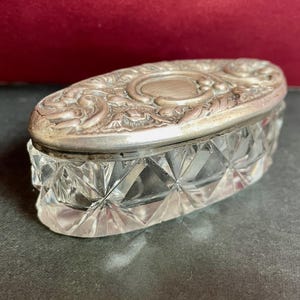 May include: An antique oval trinket box with a clear, faceted glass base and a silver-toned, ornate lid. The lid features a central oval design surrounded by floral and scrollwork patterns. The box sits on a dark grey surface.