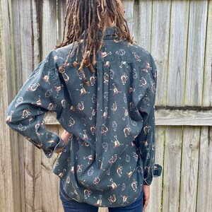 Pu&ograve; includere: Una camicia abbottonata a maniche lunghe di colore verde e blu con un motivo a forma di ferro di cavallo e altri disegni a tema equestre.