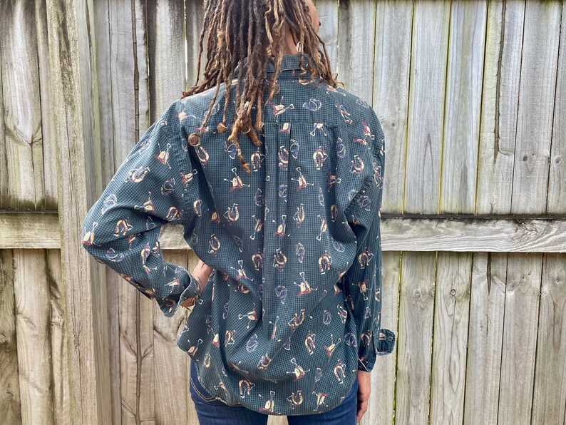 Pu&ograve; includere: Una camicia abbottonata a maniche lunghe di colore verde e blu con un motivo a forma di ferro di cavallo e altri disegni a tema equestre.