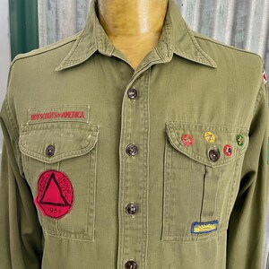 May include: Green Boy Scouts of America shirt with a red triangle patch, a yellow and green patch, and a yellow and red patch. The shirt has a pocket on each side and a button-up front.