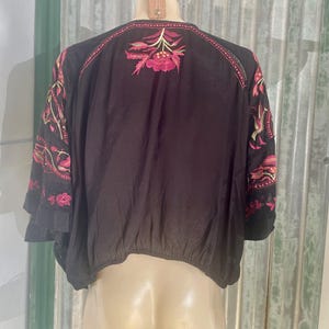 May include: Black embroidered top with pink and yellow floral designs. The top features a round neckline and three-quarter sleeves. The embroidery is concentrated on the shoulders and sleeves, creating a bohemian style.