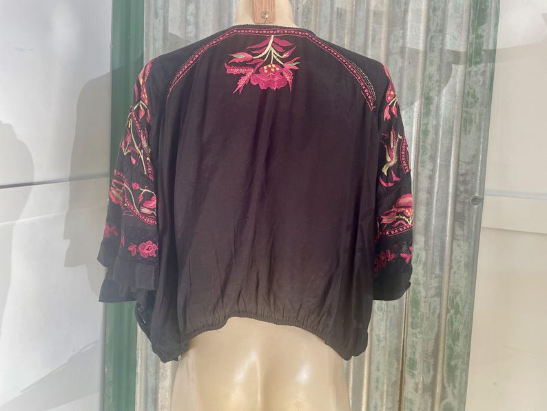 May include: Black embroidered top with pink and yellow floral designs. The top features a round neckline and three-quarter sleeves. The embroidery is concentrated on the shoulders and sleeves, creating a bohemian style.