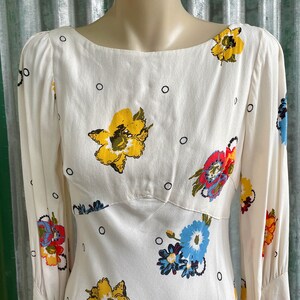 May include: A white dress with a floral print featuring yellow, red, blue, and green flowers. The dress has a scoop neck and long sleeves.
