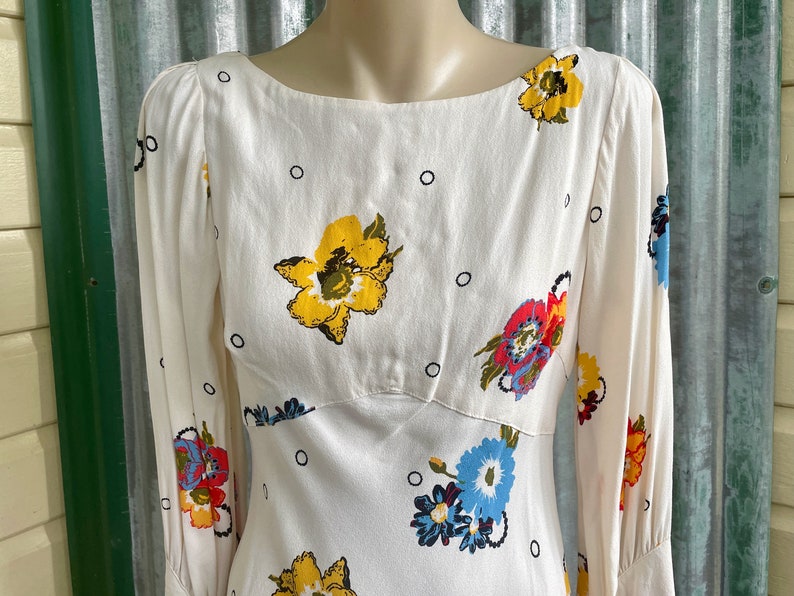 May include: A white dress with a floral print featuring yellow, red, blue, and green flowers. The dress has a scoop neck and long sleeves.