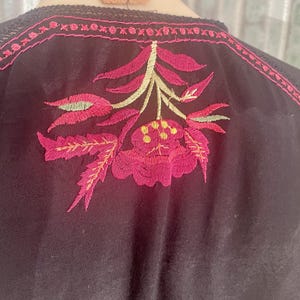 May include: Black top with intricate floral embroidery in shades of pink, green, and yellow. The embroidery is centered on the back and along the neckline, creating a detailed and colorful design. The top has a delicate, feminine aesthetic.