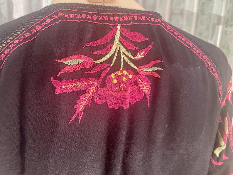 May include: Black top with intricate floral embroidery in shades of pink, green, and yellow. The embroidery is centered on the back and along the neckline, creating a detailed and colorful design. The top has a delicate, feminine aesthetic.