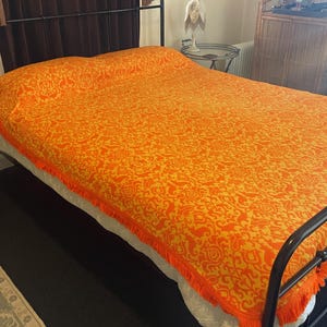 Può includere: Un copriletto arancione e giallo a fantasia con frange, che copre un letto con una struttura in metallo nero. Il copriletto presenta un motivo floreale ripetuto. Il letto è in una stanza con pavimento scuro.