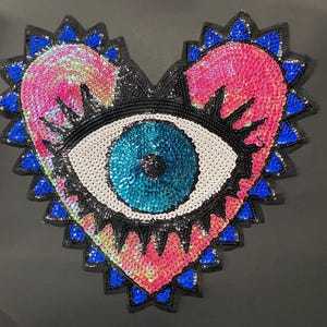 Puede incluir: Un parche de lentejuelas en forma de corazón con un diseño de ojo. El corazón está relleno de lentejuelas rosas e iridiscentes, con un borde azul y negro. El ojo tiene un iris azul, una esclerótica blanca y pestañas negras.
