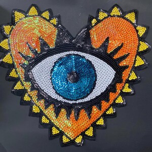 May include: A heart-shaped sequin patch featuring an eye design. The eye has a blue iris, white sclera, and black eyelashes. The heart is orange with yellow and black accents, creating a vibrant and decorative embellishment.