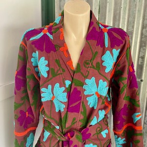 May include: A maroon robe with a floral pattern embroidered in shades of blue, green, orange, and purple. The robe has a long sleeve and a tie belt.