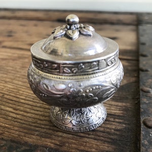 May include: A small, silver, ornate box with a lid. The box is decorated with floral patterns and has a rounded base.
