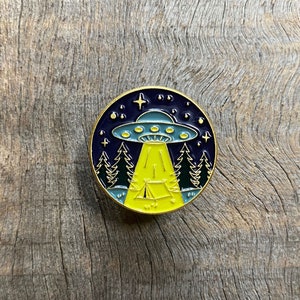 May include: A gold-colored enamel pin with a black and blue UFO hovering over a yellow tent in a forest. The UFO is emitting a yellow beam of light. The background is a dark blue night sky with stars.