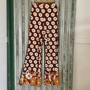 Puede incluir: Pantalón acampanado marrón con estampado de margaritas blancas. El pantalón tiene pierna ancha y cintura alta.