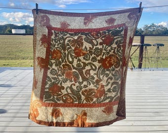 Könnte beinhalten: Ein quadratischer, transparenter Schal mit einem Blumenmuster in Rost-, Olivgrün- und Cremetönen. Der Schal hängt an einer Wäscheleine, mit einem rostfarbenen Rand. Das Design zeigt stilisierte Blumen und Paisley-Formen.