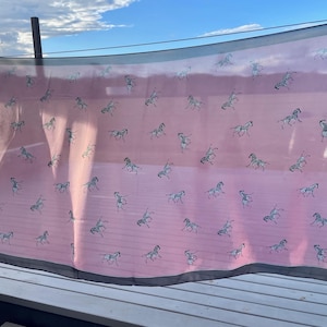 May include: A pink scarf with a repeating pattern of running horses in grey. The scarf has a dark grey border. The scarf is hanging outside on a sunny day.