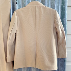 May include: A beige blazer with long sleeves and a single back vent. The blazer is made of a smooth, textured fabric.