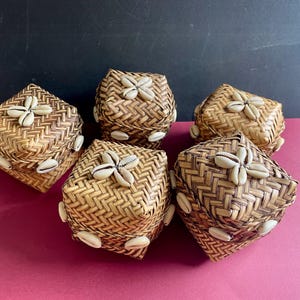 May include: Five square woven boxes with lids, adorned with seashells. The boxes are crafted from light brown woven material, presenting a natural, rustic aesthetic. Seashells are arranged on the lids and sides.