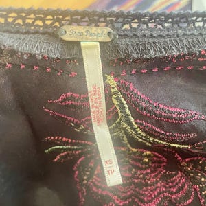 May include: Close-up of a dark gray garment with intricate embroidery in pink and green. The garment has a delicate lace trim and a tag that reads "Free People" and "Made in India."