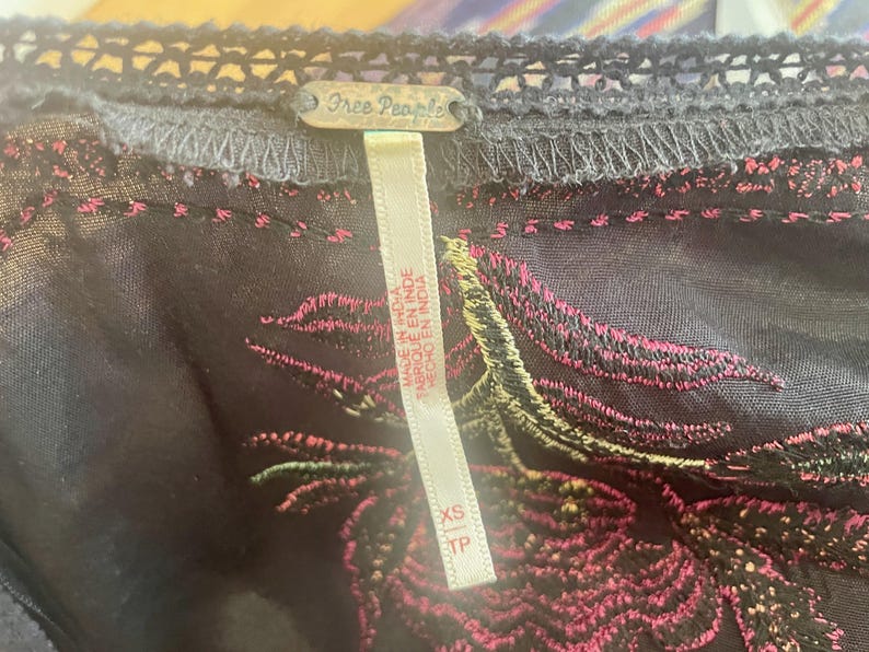 May include: Close-up of a dark gray garment with intricate embroidery in pink and green. The garment has a delicate lace trim and a tag that reads "Free People" and "Made in India."
