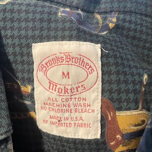 Pu&ograve; includere: Un'etichetta bianca con il testo "Brooks Brothers" in un ovale rosso, "Makers" in un ovale rosso e "M" al centro. L'etichetta include anche il testo "All Cotton, Machine Wash, No Chlorine Bleach, Made in U.S.A., Of Imported Fabric".