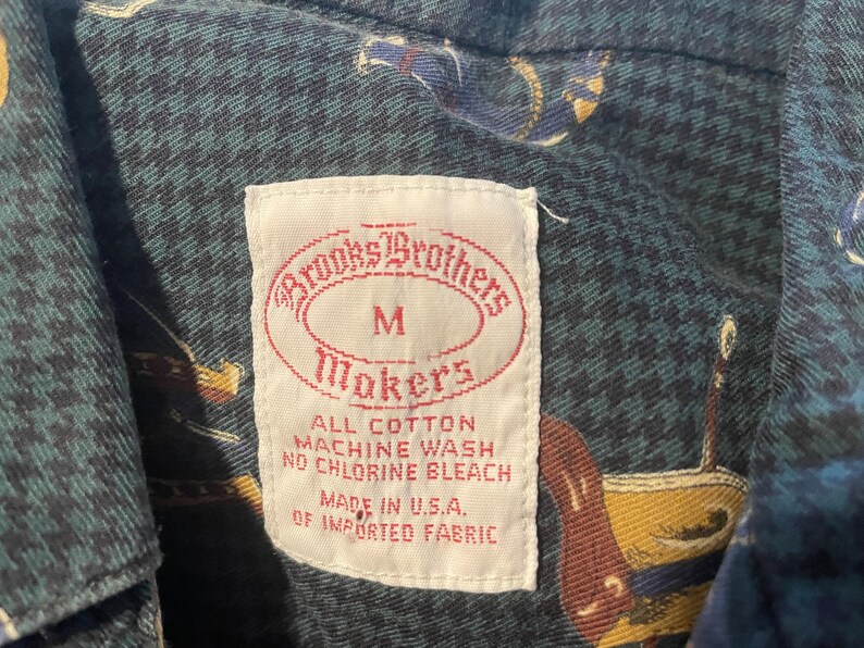 Pu&ograve; includere: Un'etichetta bianca con il testo "Brooks Brothers" in un ovale rosso, "Makers" in un ovale rosso e "M" al centro. L'etichetta include anche il testo "All Cotton, Machine Wash, No Chlorine Bleach, Made in U.S.A., Of Imported Fabric".