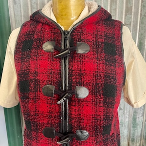 May include: A red and black plaid vest with a hood and toggle closures. The vest has a black zipper and leather-like accents. The hood has a cream-colored lining. The vest is worn over a tan short-sleeved shirt.