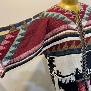 May include: A colorful open-front cardigan with a geometric pattern in red, green, blue, and yellow. The sleeves have a fringed edge. The bottom portion of the cardigan features a black and white forest scene with pockets.