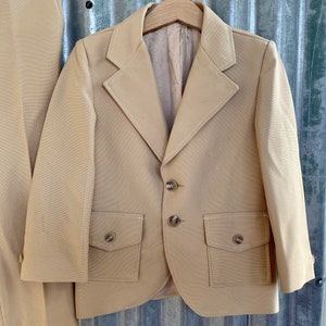 May include: A tan blazer with two flap pockets and three buttons. The blazer has a notched lapel and is made of a smooth fabric.