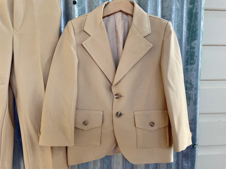 May include: A tan blazer with two flap pockets and three buttons. The blazer has a notched lapel and is made of a smooth fabric.