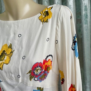 May include: White dress with a floral print featuring yellow, red, blue, and green flowers. The dress has a boat neck and 3/4 sleeves.