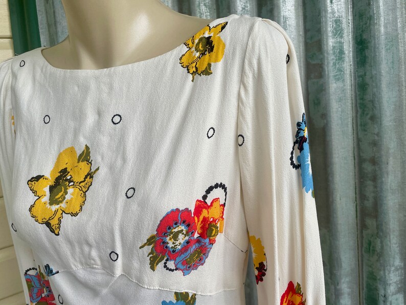 May include: White dress with a floral print featuring yellow, red, blue, and green flowers. The dress has a boat neck and 3/4 sleeves.