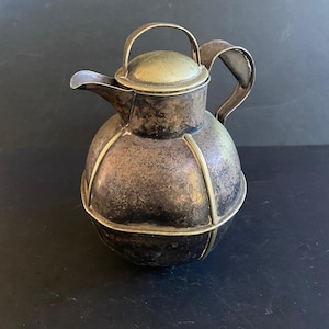 Puede incluir: Jarra antigua de metal plateado con cuerpo redondo y tapa. La jarra tiene un pico, un asa y bandas decorativas doradas. La tapa tiene un pequeño asa. Probablemente se utiliza para servir bebidas.