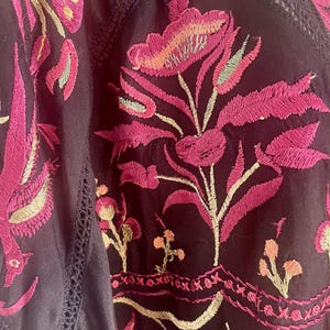 May include: Close-up of a dark purple embroidered garment. The embroidery features vibrant pink and light green floral designs. The garment has a V-neck with delicate lace trim, showcasing a bohemian style.