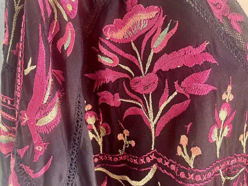 May include: Close-up of a dark purple embroidered garment. The embroidery features vibrant pink and light green floral designs. The garment has a V-neck with delicate lace trim, showcasing a bohemian style.