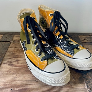 May include: A pair of high-top sneakers with a color-block design. The sneakers feature a white toe cap, black laces, and a mix of olive green, mustard yellow, and black panels. The shoes are set against a wooden surface.