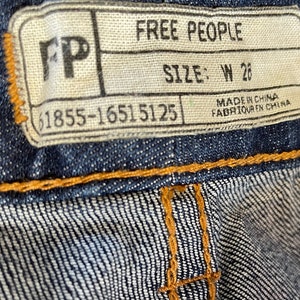 May include: A white tag with black text that reads "Free People" and "Size: W 26". The tag also includes the text "Made in China" and "Fabriqu&eacute; en Chine". The tag is attached to a pair of blue denim jeans.