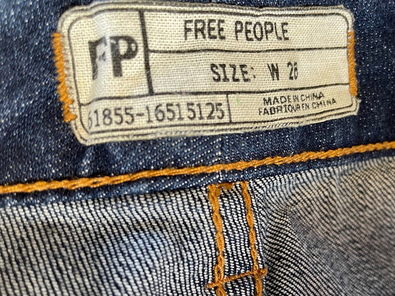 May include: A white tag with black text that reads "Free People" and "Size: W 26". The tag also includes the text "Made in China" and "Fabriqu&eacute; en Chine". The tag is attached to a pair of blue denim jeans.