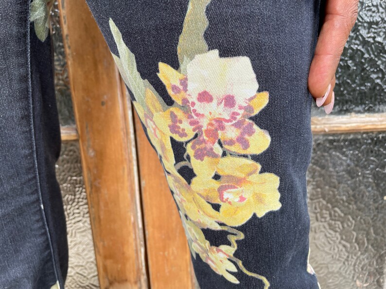 May include: Black denim pants with a floral print featuring yellow, white, and purple orchids.