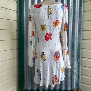 May include: A white mini dress with a floral print and a keyhole back. The dress has long sleeves and a fitted bodice.