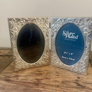 May include: Silver-plated photo frame with an oval opening. The frame is hinged and features an ornate floral design around the edges. The frame is 3.5" x 5" (9cm x 13cm) and has the words "Silver Plated PHOTO FRAME" printed on the blue backing.