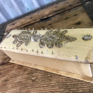 Pode incluir: Uma pequena caixa retangular coberta com tecido cor de creme com um design de contas. A caixa é decorada com um padrão de contas prateadas e brancas.