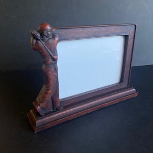 Op de afbeelding: Een donkerbruine houten fotolijst met een golferfiguur. De rechthoekige lijst heeft een leeg wit gedeelte voor een foto. De golfer is gebeeldhouwd in een zwaaiende pose, wat een sportief thema toevoegt.