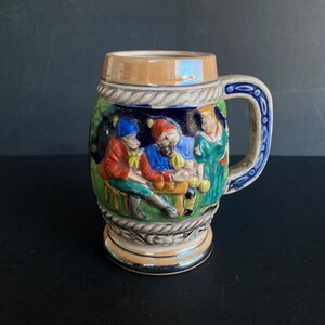 Può includere: Bicchiere da birra vintage in ceramica con manico. Il boccale presenta una scena in rilievo dettagliata di persone con abiti colorati, forse in una taverna. Il manico è blu e bianco e il bordo è marrone. Anche la base è marrone.