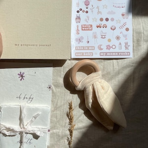 May include: A beige pregnancy journal titled GROWING YOU, a sheet of Campos & Olives baby stickers, a stack of white cards tied with ribbon labeled oh baby, and a wooden teething ring with a beige fabric attachment on a neutral fabric surface.