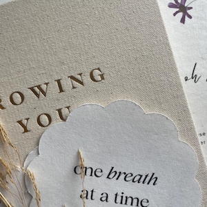 May include: A beige fabric-covered book with the gold text GROWING YOU, partially covered by a scalloped white paper card featuring the black text one breath at a time, accented by thin dried grass stems and a small purple flower illustration.