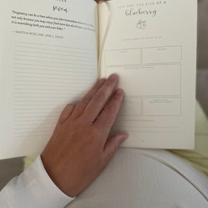 May include: An open pregnancy journal with cream-colored pages. The left page displays the text Week Seven and a quote by Martha Rose and Jane L. Davis. The right page features the text You are the size of a blueberry with a small illustration.
