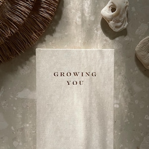 May include: A beige journal with the words "GROWING YOU" embossed on the cover. The text "my pregnancy journal" is printed below. The journal is lying on a textured surface, with decorative elements in the background.