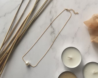 May include: A delicate gold chain necklace with a single pearl pendant. The necklace is displayed on a white marble surface with wheat stalks and candles.