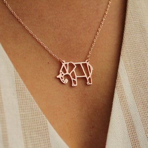 May include: Rose gold elephant pendant necklace. The pendant is a geometric design of an elephant. The necklace is on a delicate chain. The background is a cream-colored fabric.