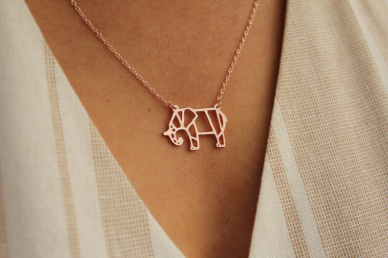 May include: Rose gold elephant pendant necklace. The pendant is a geometric design of an elephant. The necklace is on a delicate chain. The background is a cream-colored fabric.